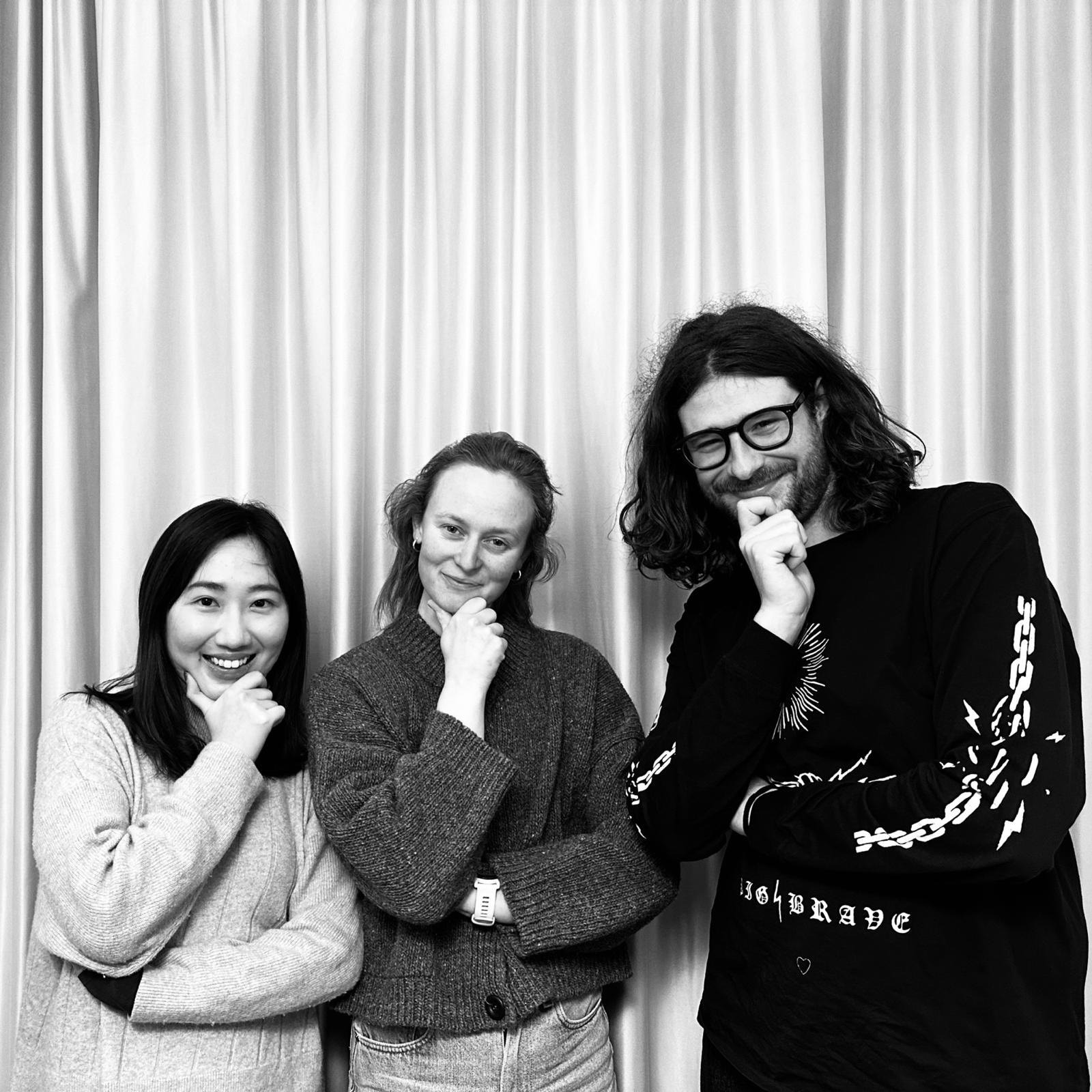 This is a black-and-white portrait featuring three individuals—two women and one man—standing side-by-side against a backdrop of light-colored, vertically pleated curtains.

The subjects are posed in a way that suggests a shared, playful, yet intellectual theme: all three are looking toward the camera with slight smiles, each resting their chin on one hand in a "thinking" gesture.

    On the left is a young woman with dark, straight hair and a bright, wide smile. She is wearing a light-colored, textured knit sweater.
    In the center is a woman with lighter hair pulled back, wearing a subtle smile. She is dressed in a dark, chunky, textured cardigan over jeans and is wearing a watch on her left wrist.
    On the right is a man with dark, wavy, shoulder-length hair, a beard, and glasses. He has a knowing smirk and is wearing a dark, long-sleeved graphic t-shirt. The shirt features white designs, including a chain and lightning bolt pattern on the sleeve and stylized text on the chest that includes the word "BRAVE."

The overall composition is clean and centered, with the black-and-white medium emphasizing the textures of their clothing and the vertical lines of the background curtains. Given the provided context, the image serves as a professional yet approachable group portrait of the researchers Charmaine Kong, Laura Wohlgemuth, and Alessandro Pellanda.